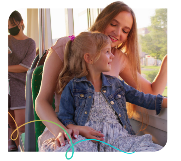 Side view of woman holds her daughter on her lap in streetcar bus. Happy family rides in public transport train, mom with little blonde girl sit together and look out window tram, talking, laughing